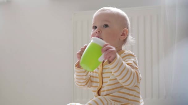 Portrait drinking baby at home. Child learns to drink on his own. Kid sitting on floor with plastic green cup in hands . Healthy food, lunch time — Stock Video