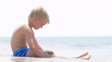 Kumsaldaki okyanusta kumla oynayan küçük sarışın çocuk. Aile tatilleri ve sıcak ülkelere seyahatler. Mutlu turizm tatili Mavi dalgalar suyu.