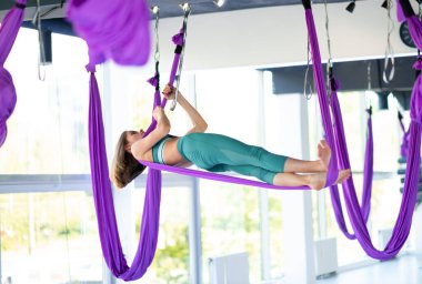 Genç, gülümseyen kadın salıncakta sallanıyor. Havadan uçan yoga egzersizleri spor salonunda mor hamakta pratik yapıyor.