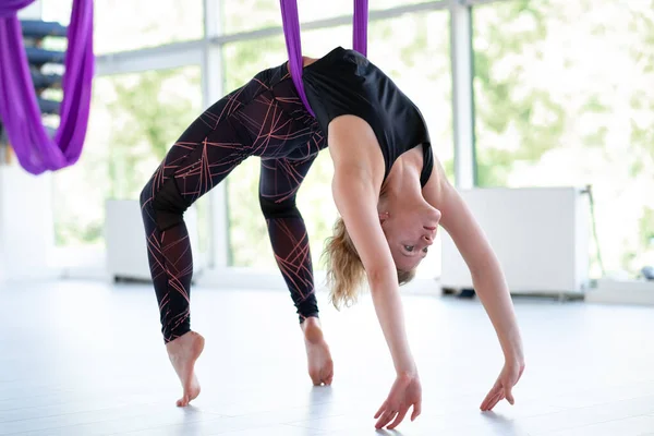 Young smiling woman practice in aero stretching swing. Aerial flying yoga exercises practice in purple hammock in fitness club