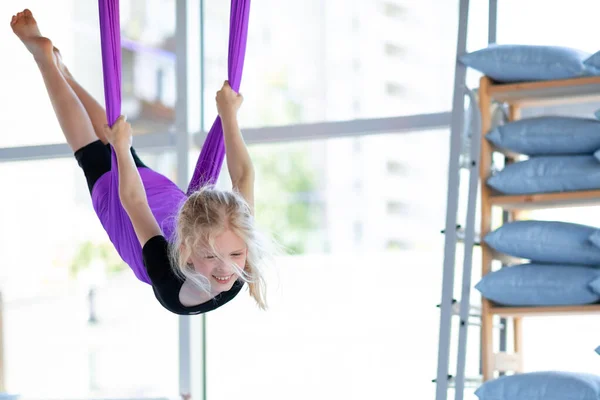 Havacılık antrenmanında gülümseyen genç bir kız spor salonunda mor hamakta salıncak sallıyor. Çocuklar hava uçuş yoga egzersizleri.