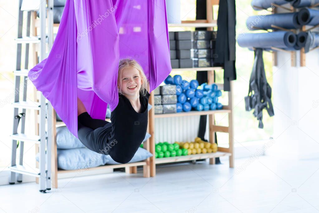 Portrait young smiling girl practice in aero stretching swing in purple hammock in fitness club. kids Aerial flying yoga exercises
