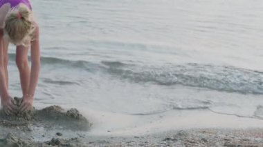 Gün batımında kumsalda kumla oynayan küçük sarışın kız. Çocuk kumdan kale inşa ediyor. Aile yaz tatilleri ve sıcak ülkelere seyahatler.