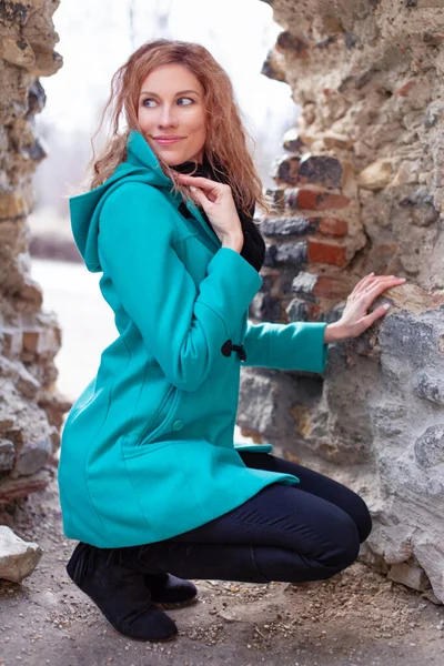 Sexy young redhead woman squats at ruins in long turquoise coat, looking away