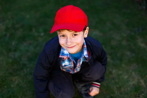 Menino em chapéu vermelho agachamento ao ar livre na grama — Fotografia de Stock