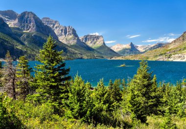 Buzul Ulusal Parkı 'ndaki Vahşi Kaz Adası' nın canlı mavi Saint Mary Gölü 'nün panoramik görüntüsü. Rocky Dağları, çam ağaçları tarafından çerçevelenmiş berrak mavi bir gökyüzünün altında görkemli bir şekilde yükselir.