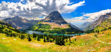 Montana 'daki Buzul Ulusal Parkı' ndaki Logan Geçidi 'nden Saklı Göl ve Bearhat Dağı' nın geniş panoramik manzarası. Yeşil çayır, dramatik bulutlu bir gökyüzünün altında ön planda.