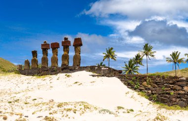Paskalya Adası 'ndaki Anakena Sahili' nde Ahu Nau Nau üzerinde kırmızı pukao topuzları olan Moai heykelleri duruyor. Beyaz mercan kumu ve palmiye ağaçları ön plandadır.