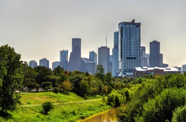 Houston şehir merkezinin ufuk çizgisi yemyeşil ağaçların ve Buffalo Bayou Parkı 'nın üzerinde yükseliyor. Çamurlu kahverengi bataklık ön planda akıyor.