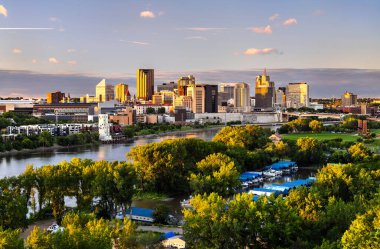 Saint Paul şehir merkezi ABD 'nin Minnesota kentindeki Mississippi Nehri üzerinde yükselir. Manzarada şehir mimarisi ve günbatımı altında yeşil ağaçlar yer alıyor.