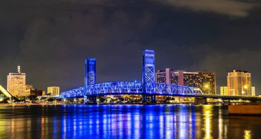 Jacksonville Florida 'da geceleyin ana cadde köprüsü. Tarihsel mavi ışıklı dikey kaldırma köprüsü arka planda aydınlık şehir silueti ile St. Johns Nehri boyunca uzanır.