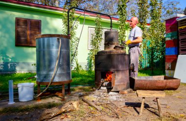 Man is manually turning lever to mix fruit marc in boiler of homemade distillery made of copper, making moonshine schnapps, alcoholic beverages such as brandy, cognac, whiskey, bourbon, gin, scotch. clipart