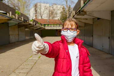 Tıbbi maske ve eldiven takan küçük bir kız COVID-19 virüsünden korunmak için baş parmağını kaldırıyor..