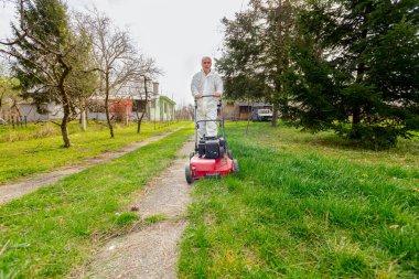 Koruyucu giysili bahçıvan bahçesinde çimen biçiyor. Çim biçme makinesi de meyve ağaçları arasında..