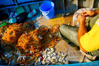 Açık denizde balık tutmak için teknesinde oturup balık ağlarını üst üste yığan lastik pantolonlu balıkçı..