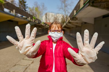 Tıbbi maske takan ve eldiven takan küçük kız ellerini, jestlerini, sosyal mesafelerini, uzak durduğunu, COVID-19 virüsüne karşı korunduğunu gösteriyor..