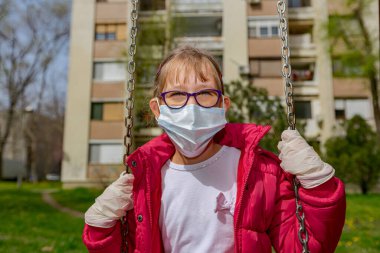 Tıbbi maskeli ve eldivenli küçük kız COVID-19 virüsünden korunmak için tek başına sallanıyor.. 