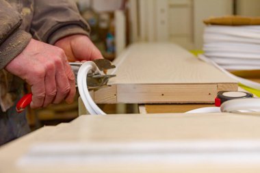 Marangoz işçisi elinde kırmızı metal pense tutuyor ve marangozhanedeki ahşap kapı çerçevesine yerleştirilmiş plastik bant kesiyor..