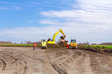 Proje devam ediyor, kazıcı çöp kamyonunu toprakla dolduruyor ve yol silindiri inşaat alanını düzleştiriyor, düzleştiriyor, eşitliyor.