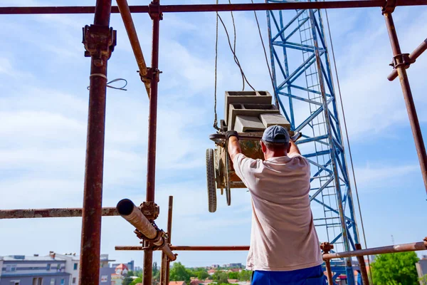 Worker is helping crane to keep balance and direction as transport wheelbarrow laden with blocks on scaffold with wooden platform building site.