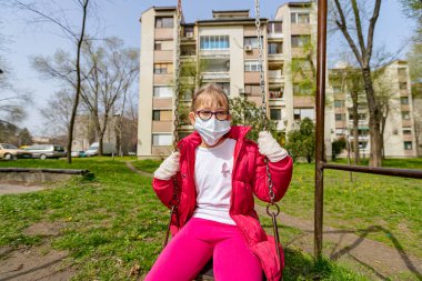 Tıbbi maskeli ve eldivenli küçük kız COVID-19 virüsünden korunmak için tek başına sallanıyor.. 