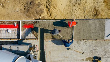 Üst manzarada, inşaat işçilerinin genel giderleri tırmıkla yayılıyor ve mikser kamyonundan dökülen taze betonu kare siperlere yerleştirmek için metal katman kullanılıyor..