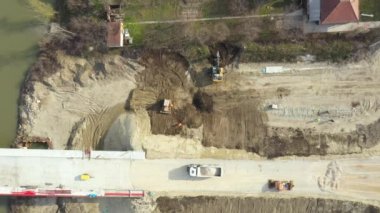 Üst manzarada, tepede, tamamlanmamış beton köprüde nehrin üzerindeki yol yapım aşamasında. Kazıcı inşaat alanında toprak çıkarıyor. Proje devam ediyor..