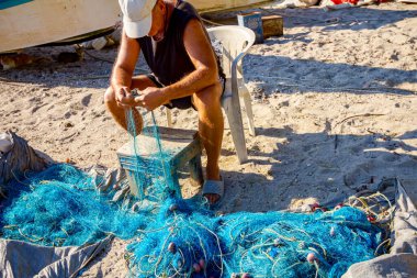 Bir balıkçı sahilde bir sandalyede oturur, balık ağını biriktirir ve kabuklarından temizler..