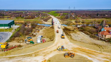 Manzaranın üstünde, dairesel yolda, inşaat halindeki döner kavşakta, inşaat alanındaki işçiler ağır makinelerle yol temeli üzerinde çalışıyorlar..