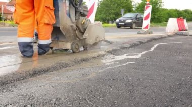 İşçi, asfaltta düzgün bir kesik yapmak için dairesel elmas testeresi kullanıyor. Testere yüksek hızda döndüğü için malzemeyi kesmek için aşındırıcı bir etki kullanıyor..