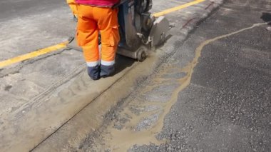 İşçi, asfaltta düzgün bir kesik yapmak için dairesel elmas testeresi kullanıyor. Testere yüksek hızda döndüğü için malzemeyi kesmek için aşındırıcı bir etki kullanıyor..