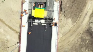 Above dolly move, top view, overhead, on workers and machinery for laying asphalt, spreading layer of hot tarmac over unfinished bridge, road over river is under construction, building site.