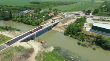 Above dolly move, sideways on workers and machinery for laying asphalt, spreading layer of hot tarmac over new bridge, road over river is under construction, building site. Circular road, roundabout
