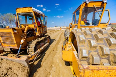 Dikenli ve buldozerli yol silindiri, inşaat alanındaki yol temeli için kum seviyesini düşürüyor..