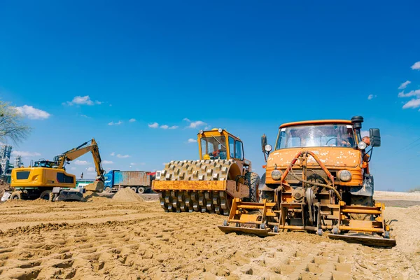 Road roller with spikes and truck with mounted plate vibration compactor are compacting, leveling sand for road foundation at building site.