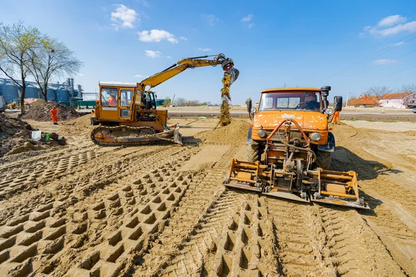 View on excavator and truck with mounted plate vibration compactor, as they are compacting, leveling sand for road foundation at building site, mechanization.