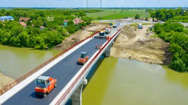 Yukarıda, asfalt döşeyen işçiler ve makinelerde, yeni köprünün üzerine sıcak asfalt tabakası yayılıyor, nehrin üzerindeki yol yapım aşamasında. Dairesel yol, arka planda trafik olan döner kavşak.