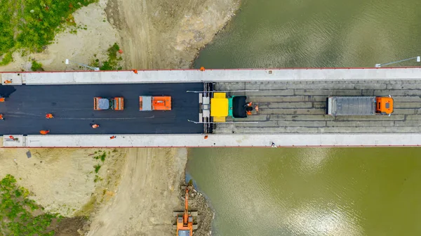 上記のビュー オーバーヘッドは 未完成の橋 アスファルトを敷設するための労働者や機械の上に熱いTarmacのトラック配信と輸送 川の上に道路層を広げる — ストック写真