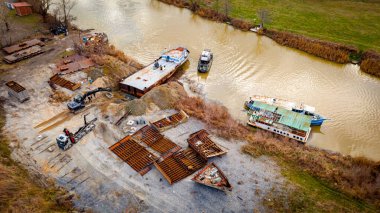 Above view over excavator, tow truck and tugboat as they pull out part of old freight vessel, barge, at river coast, cassation, dismantling ship, metal for scarp. Workers cutting into smaller pieces