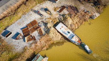 Above view over excavator, tow truck and tugboat as they pull out part of old freight vessel, barge, at river coast, cassation, dismantling ship, metal for scarp. Workers cutting into smaller pieces