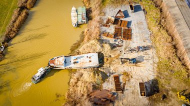 Above view over excavator, tow truck and tugboat as they pull out part of old freight vessel, barge, at river coast, cassation, dismantling ship, metal for scarp. Workers cutting into smaller pieces