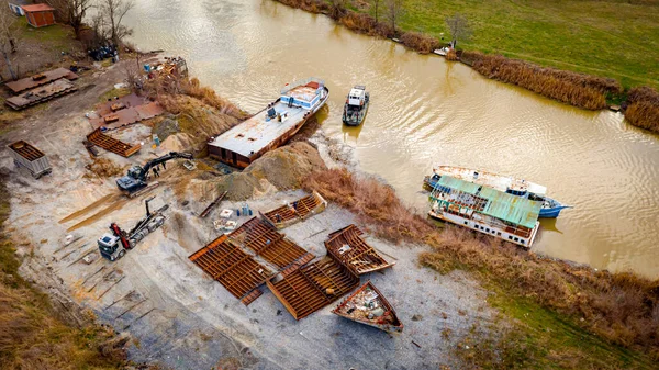 Above view over excavator, tow truck and tugboat as they pull out part of old freight vessel, barge, at river coast, cassation, dismantling ship, metal for scarp. Workers cutting into smaller pieces