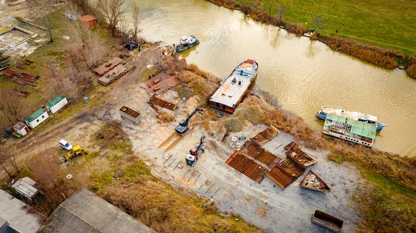 Above view over excavator, tow truck and tugboat as they pull out part of old freight vessel, barge, at river coast, cassation, dismantling ship, metal for scarp. Workers cutting into smaller pieces