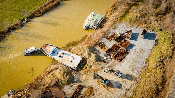 Above view over excavator, tow truck and tugboat as they pull out part of old freight vessel, barge, at river coast, cassation, dismantling ship, metal for scarp. Workers cutting into smaller pieces