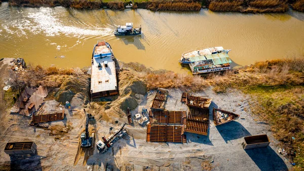 Above view over excavator, tow truck and tugboat as they pull out part of old freight vessel, barge, at river coast, cassation, dismantling ship, metal for scarp. Workers cutting into smaller pieces