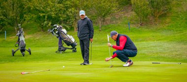 İnsanlar golf oynuyor, golf oynarken çimlerin üzerinde yürüyorlar. İçinde ekipman ya da yük arabası olan çanta taşıyorlar..