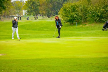 İnsanlar golf oynuyor, golf oynarken çimlerin üzerinde yürüyorlar. İçinde ekipman ya da yük arabası olan çanta taşıyorlar..