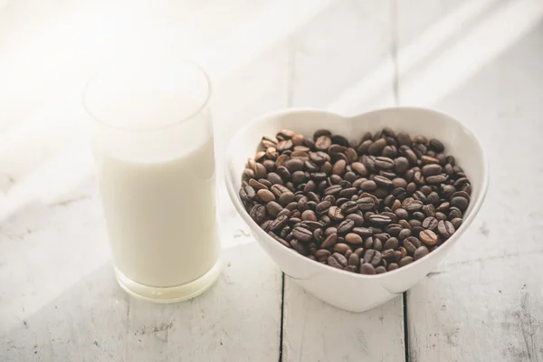 Kávová zrna v misce a sklenici mléka na dřevěný stůl. — Stock fotografie
