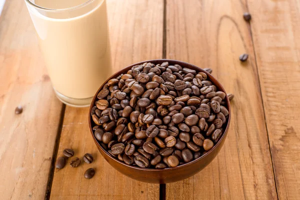 Coffee beans in bowl and a glass of coffee milk on wooden table. — Stock Photo, Image