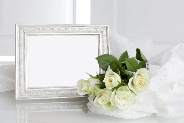Leerer silberner Bilderrahmen und fünf weiße Rosen auf weißer Spitze Stockbild